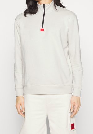 Henkilö pukeutuneena off-white-väriseen puolivetoketjulliseen svetariin ja off-white-värisiin housuihin, joissa on sivutasku; molemmissa pienet punaiset logoplaasit.