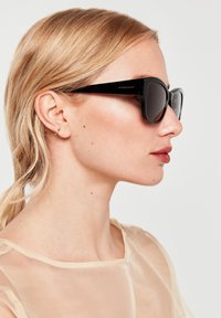 Gafas de sol rectangulares negras con montura gruesa, lentes oscuros y acabado brillante. Acompañadas de una blusa transparente de color claro.