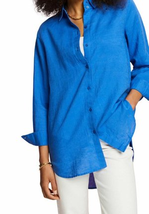 Femme portant une chemise bleue ample boutonnée avec des poignets retroussés et un pantalon blanc, main dans la poche, sur un fond blanc uni.