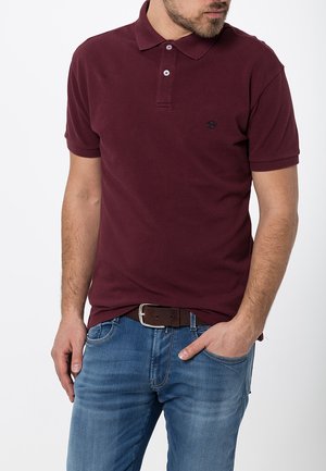 Bordeaux Kurzarm-Polo-Shirt mit Kragen und drei Knöpfen; aus Baumwollstoff; mit einem kleinen Logo auf der linken Brust. Getragen mit blauen Jeans.