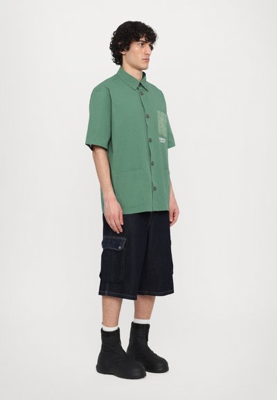 Zelena kratka majica s kratkimi rokavi in žepom, v kombinaciji s temnimi denimskimi kratkimi hlačami. Model nosi črne škornje na zadrgo in bele nogavice.