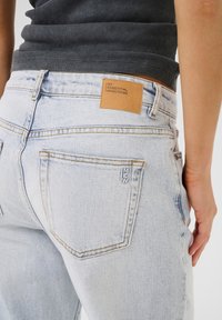 Svijetloplave traper hlače s kožnim detaljem na pojasu s utisnutim tekstom. Vidljivi istrošeni detalji i standardni džepovi.