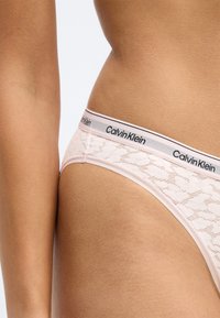 Calvin Kleini roosa pitsiline stringkandja pehme tekstuuriga, millel on kontrastne must logoga vöökoht ja lilleline muster üle kogu toote.