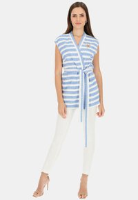 Niebieska i biała w paski bezrękawnikowa bluzka w fasonie kopertowym z paskiem, wykonana z teksturowanej tkaniny. Posiada dekoracyjną szpilkę w okolicy klatki piersiowej.