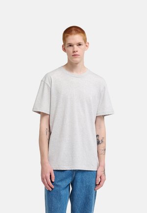 Jonge man met kort rood haar draagt een lichtgrijs T-shirt en blauwe spijkerbroek, staat met een neutrale uitdrukking en zichtbare tatoeages op de onderarmen.