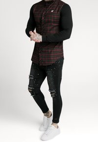Camicia a quadri rossa e nera con maniche lunghe nere, abbinata a jeans neri usurati con accenti di vernice colorata e sneakers bianche.