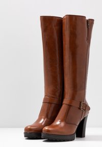 Bottes montantes en cuir marron avec un bout arrondi, un accent boucle et un talon bloc. Texture lisse avec une fermeture éclair latérale.