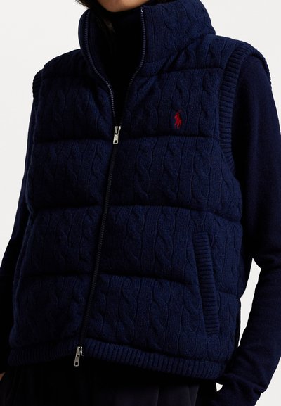 Puffa prsluk u tamnoplavoj boji s rebrastom teksturom, visokim ovratnikom, zatvaranjem na zip, i bočnim džepovima. Crveni logotip na prsima.