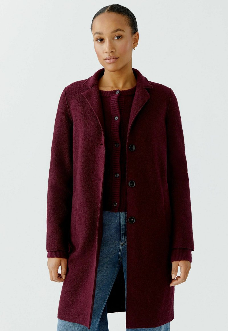 Burgundy wool-blend coat with a notched collar, long sleeves, and black buttons, layered over a ribbed burgundy sweater and denim jeans.