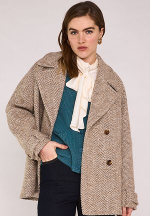 Jonge vrouw met beige visgraatjas, teal cardigan, crème sjaal, gouden oorringen en donkere broek, hand in zak, neutrale achtergrond.