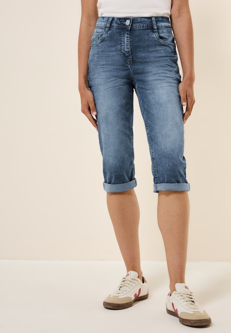 Pantalones capri de mezclilla en azul claro con puños enrollados, con una cintura de altura media y bolsillos delanteros y traseros. Llevados con zapatillas blancas.