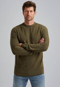 Olivovo zelený pletený sveter s rebrovanou textúrou, okrúhlym výstrihom a malou emblémou na hrudi, skombinovaný so svetlomodrými džínsami.