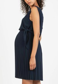 Abito plissettato blu navy con corpetto aderente, dettaglio di fiocco laterale e bottoni decorative sulla spalla. Realizzato in un morbido tessuto testurizzato.