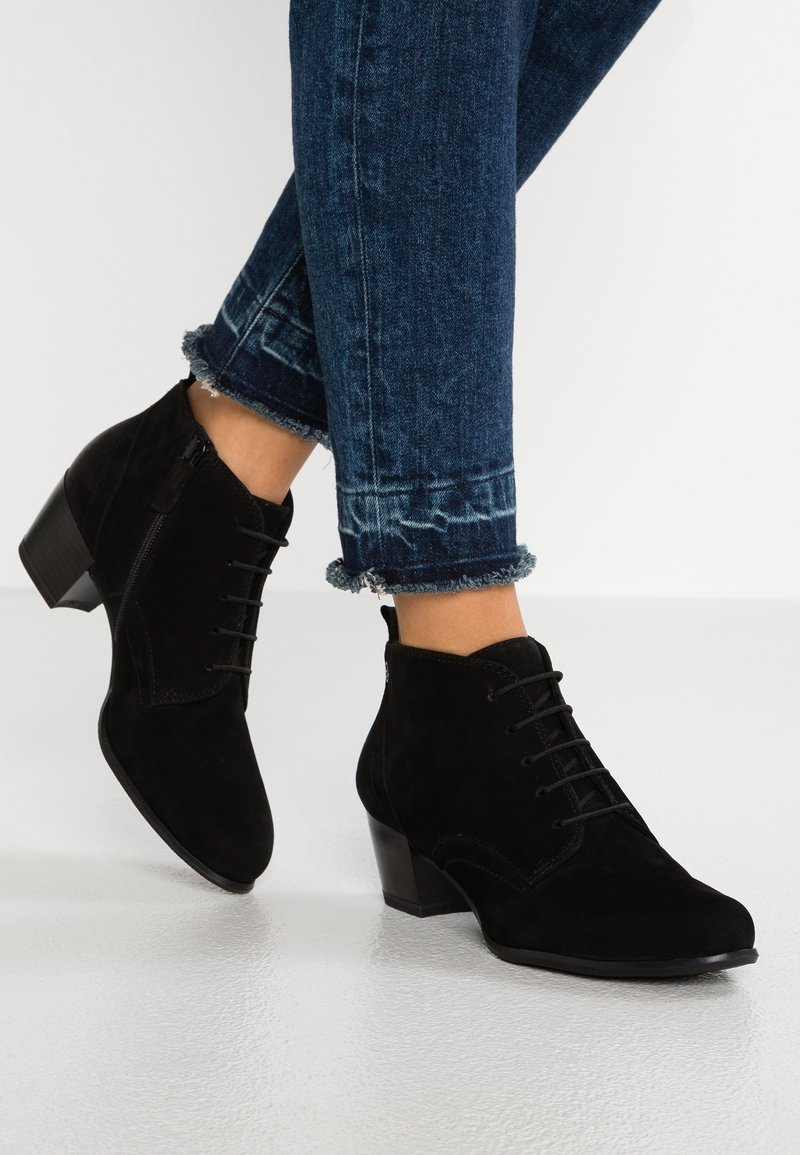 Botas de ante negras hasta el tobillo con cordones en la parte delantera, punta redondeada y tacón medio. Llevadas con unos vaqueros azules ligeramente desgastados y acortados.