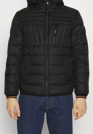 Mies pukeutuneena mustaan tikattuun untuvatakkiin, jossa on etuvetoketju, vetoketjullinen rintatasku ja painonappikiinnitteiset sivutaskut, yhdistettynä sinisiin farkkuihin.