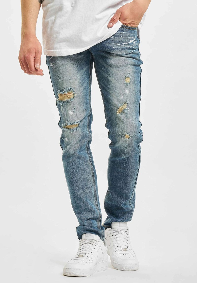 Bleket blå jeans med flere slitte områder og lapper. Bært med en hvit t-skjorte og hvite sneakers, med en slank passform.