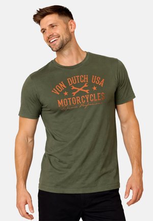 Homme souriant portant un T-shirt vert olive avec le texte orange « Von Dutch USA Motorcycles » et un graphisme de clé, debout devant un fond uni.