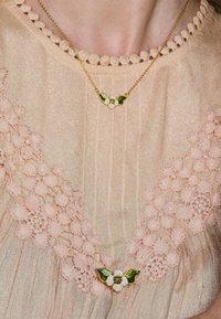 Goldene Halskette mit floralem Design, weißen Blütenblättern und grünen Blättern, getragen über einem beigefarbenen, strukturierten Kleidungsstück mit pinkfarbenen Akzenten.