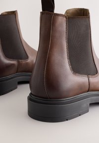 Hnedé kožené chelsea topánky s elastickými bočnými panelmi, štruktúrovaným povrchom a čiernou gumovou podrážkou. Majú pútko na jednoduchšie obúvanie.