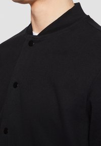 Veste noire avec col mandarin, fermetures à boutons-pression et tissu texturé. Détails visibles aux épaules et sur les coutures latérales.