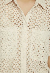 Camisa de encaje en color blanco roto con un patrón de cuadrícula, que presenta un cierre de botones y dos bolsillos en el pecho. Textura suave y diseño ligero.