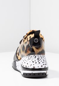 Baskets avec un tissu à imprimé léopard, détails en vernis noir et semelle blanche avec des taches noires, présentées de dos sur une surface blanche.