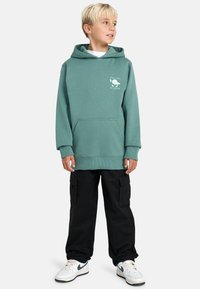 Pojke som bär en turkossfärgad huvtröja med texten "CHILL OUT. BE WATER", svarta cargo-byxor och vita sneakers med grön swoosh, står och tittar åt höger.