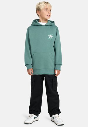 Jongen in een teal hoodie met de tekst "KALM AAN. WEES WATER", zwarte cargobroek en witte sneakers met groene swoosh, staande en kijkend naar rechts.