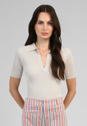 Femme aux longs cheveux foncés portant un haut beige côtelé à manches courtes et un pantalon taille haute à rayures verticales rouges, bleues et blanches.