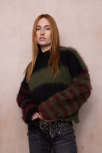 Junge Frau mit langen roten Haaren, die einen flauschigen gestreiften Pullover und dunkle Jeans mit einem gepunkteten schwarzen Gürtel trägt, steht vor einer blassrosa Wand.