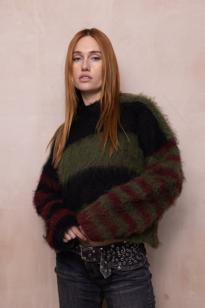 Junge Frau mit langen roten Haaren, die einen flauschigen gestreiften Pullover und dunkle Jeans mit einem gepunkteten schwarzen Gürtel trägt, steht vor einer blassrosa Wand.