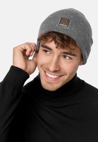 Gray knit beanie with ribbed texture and small square patch featuring logo. Worn on model, paired with black high-neck sweater.