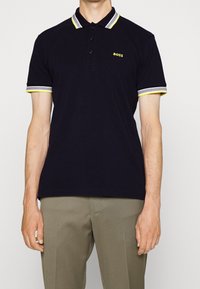 Polo shirt bleu marine avec un plastron à trois boutons, col et poignets rayés jaunes et gris contrastants, et logo "BOSS" en jaune.