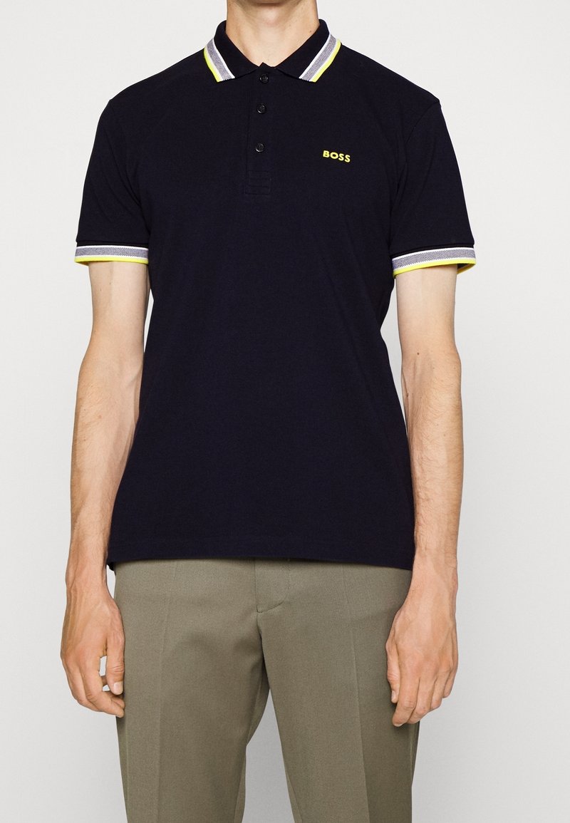 Polo shirt bleu marine avec un plastron à trois boutons, col et poignets rayés jaunes et gris contrastants, et logo "BOSS" en jaune.
