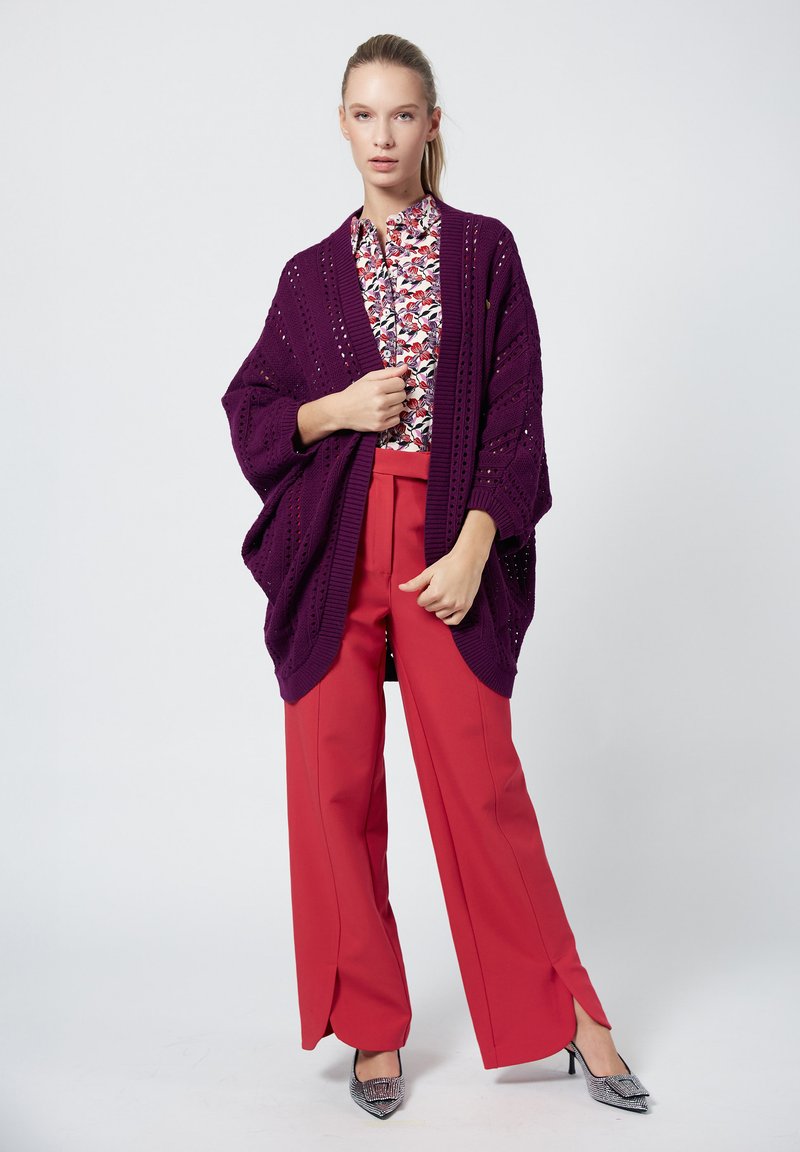 Gebreide paarse open cardigan met grote gaasdetails, gedragen over een bloemenblouse en rode wijde broek, aangevuld met zilveren hoge hakken.