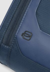 Bolso azul con textura que presenta un material liso, cierre de cremallera y un acento de logotipo negro. El diseño es elegante con un contraste de colores distintivo.