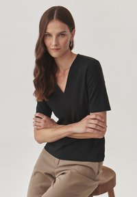Haut noir à manches courtes en V en tissu lisse, associé à un pantalon beige taille haute. Design minimaliste et silhouette ajustée.