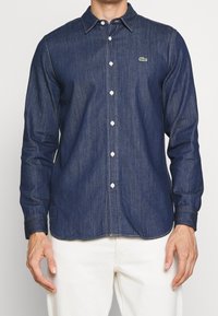 Man som bär en välpassande mörkblå denimskjorta med vita knappar och en liten grön logotyp, matchad med vita byxor.