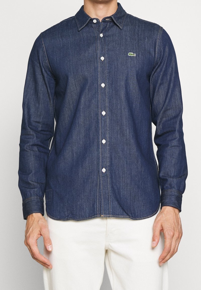 Man som bär en välpassande mörkblå denimskjorta med vita knappar och en liten grön logotyp, matchad med vita byxor.