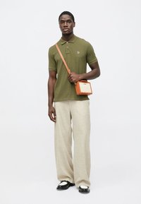 Olivzaļš polo krekls ar īsām piedurknēm, gaisīgi bēšas platās bikses un neliela oranža krūšu soma ar teksturētu rakstu. Melnas kurpes ar baltām detaļām.