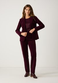 Burgundy velvet suit with tailored blazer and slim-fit trousers. The blazer features a single button, worn over a matching top and complemented by ankle boots.