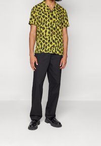 Camicia a maniche corte con un motivo leopardato verde e nero, abbinata a pantaloni neri e scarpe nere. Presenta una vestibilità rilassata e chiusura a pulsante.
