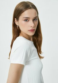 Hvid ribbet T-shirt med korte ærmer, der har en tætsiddende form og glat tekstur. Modellen viser en sidevinkel, med hår ned. Enkelt baggrund.