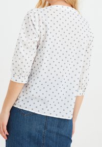 Blusa bianca con stampa floreale blu navy, tessuto leggero, maniche a tre quarti e dettagli con bottoni sui polsini, abbinata a una gonna di jeans.