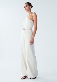 Weißes, geripptes One-Shoulder-Top kombiniert mit hoch taillierter, weiter beige Hose. Die Textur ist glatt mit einer maßgeschneiderten Passform und dezenten Nahtdetails.