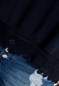 Svart topp med spetsdetalj vid fållen. Tyget är mjukt med en lätt lyster. Bärs över slitna jeans med synliga knappar och fickor.