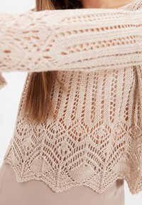 Beżowy sweter z dzianiny o wzorze koronkowym, z otwartym splotem i falbaniastym dołem, podkreślający fakturę oraz skomplikowany design.
