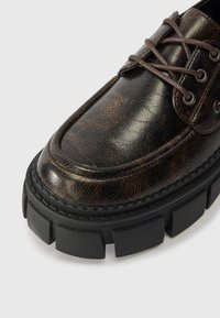 Hnědé opotřebované kožené boty s kulatou špičkou, kontrastními tmavými tkaničkami a robustní černou gumovou podrážkou s výraznými detaily dezénu.