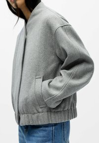 Veste bomber en polaire grise avec un col haut, des poches latérales et un ourlet côtelé. Elle présente une coupe décontractée et une texture subtile.
