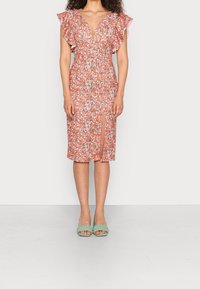 Robe fleurie orange et blanche, avec des manches volantées, un décolleté en V profond et une fente à l'avant. Assortie à des sandales à talons tressées vertes.